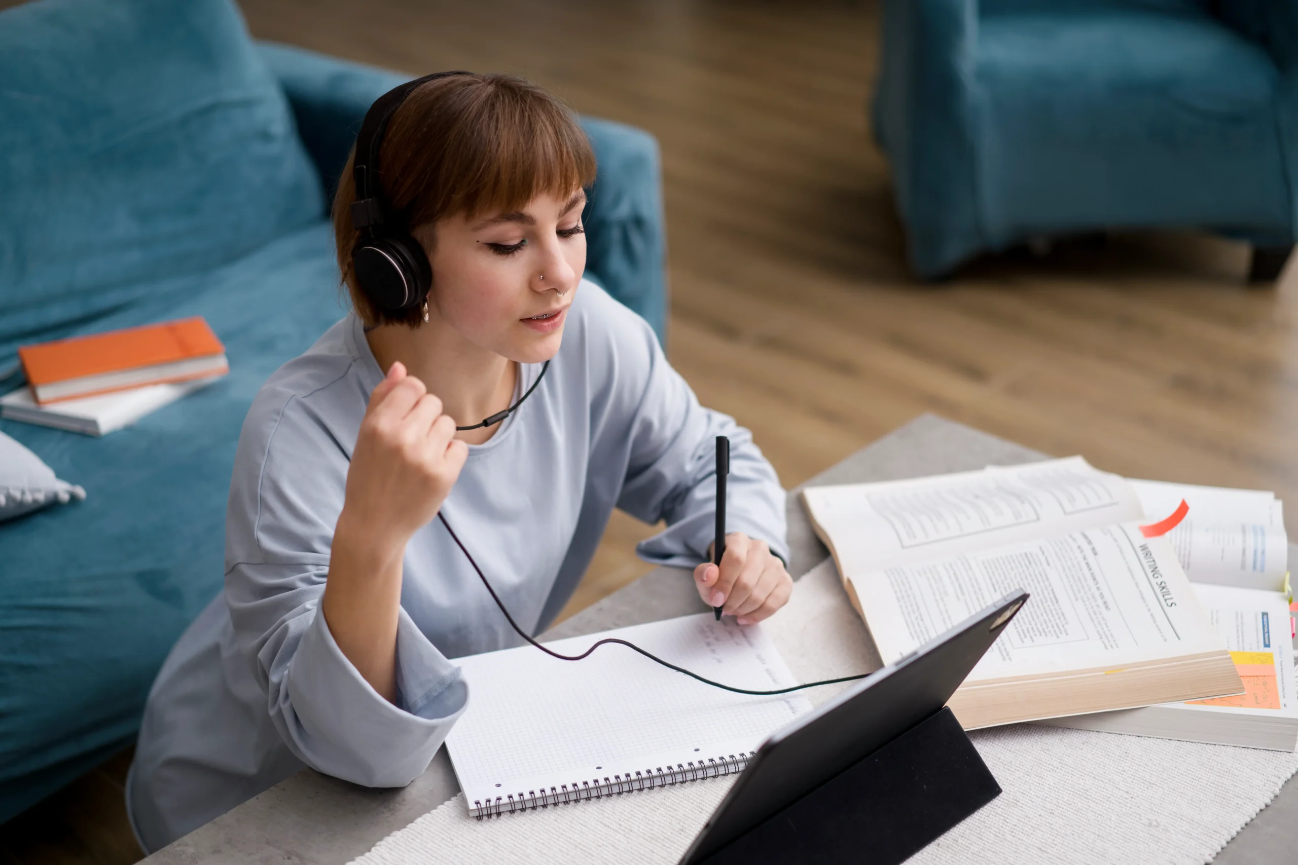 Mujer sentada en el suelo tomando notas en un cuaderno frente a una tablet, usando auriculares, mientras realiza un programa online